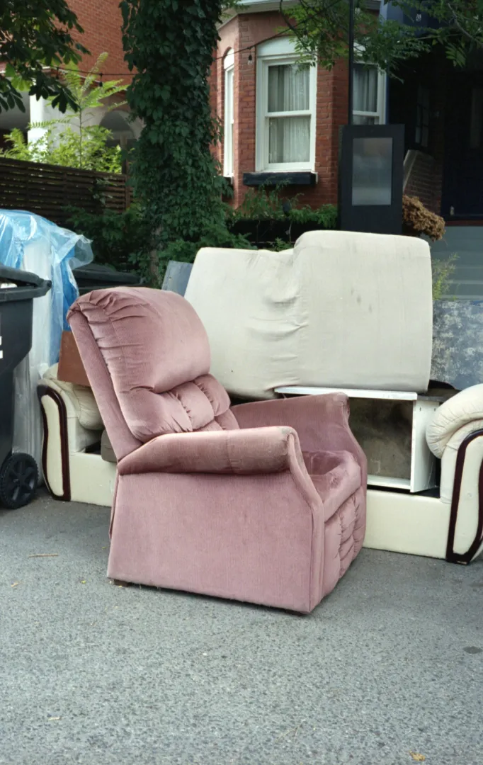 Pink recliner and cream sofa pieces set out on the street next to a trash cart in front of brick row houses