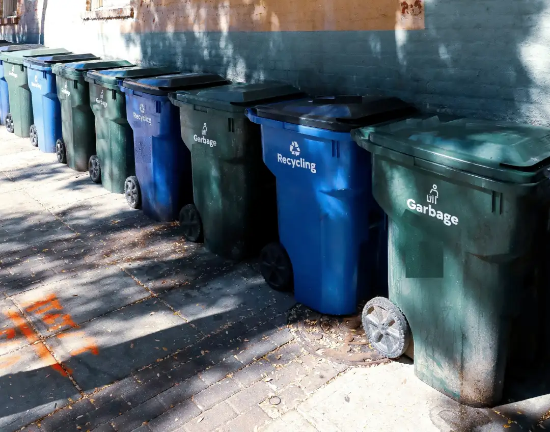 How to Schedule Waste Disposal:A row of green garbage carts and blue recycling carts lined up along a brick building wall