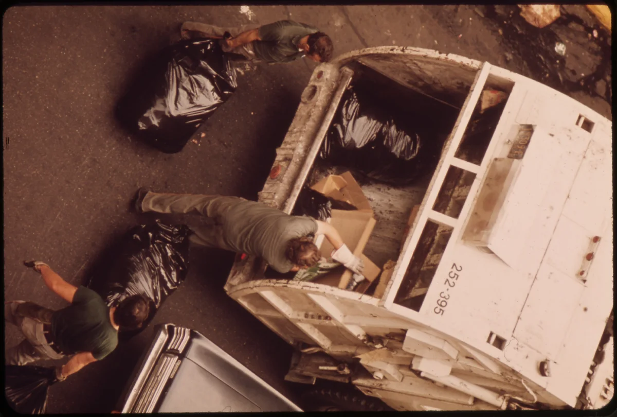 Sanitation Workers Loading a Rear-Loader Collection Truck; Manhattan Industrial Trash Compactor: Sanitation workers lifting garbage bags and a cardboard box into the open hopper of a rear-loader collection truck on a Manhattan street
