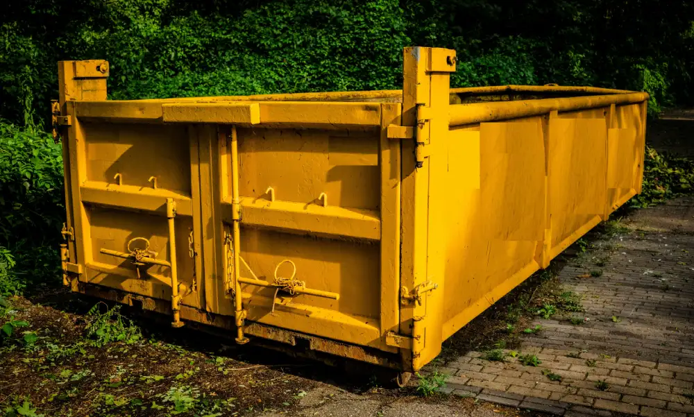 Dumpster Truck: Empty yellow roll off container set down at the edge of a road, hook plate at the front and rollers at the rear